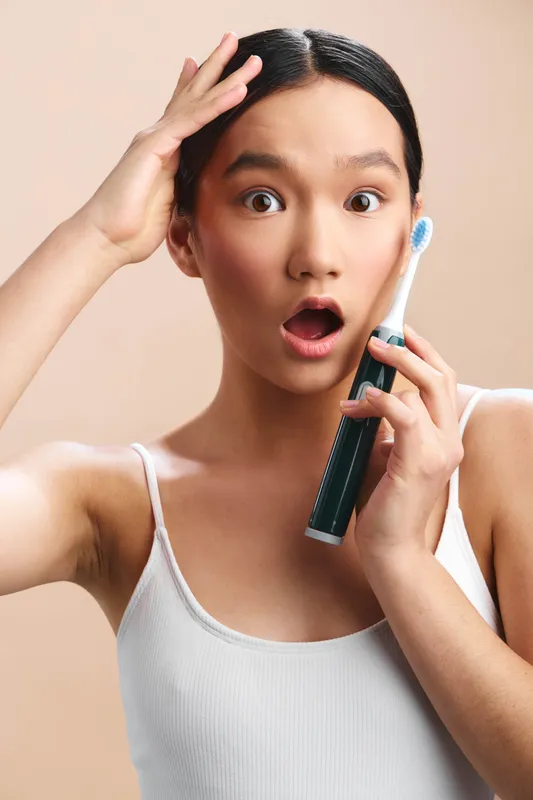 asian woman brushing teeth in a bathroom, surprised expression, wide eyes, holding toothbrush near mouth, morning routine vibe, clean fresh atmosphere, neutral backdrop, everyday wellness and care.