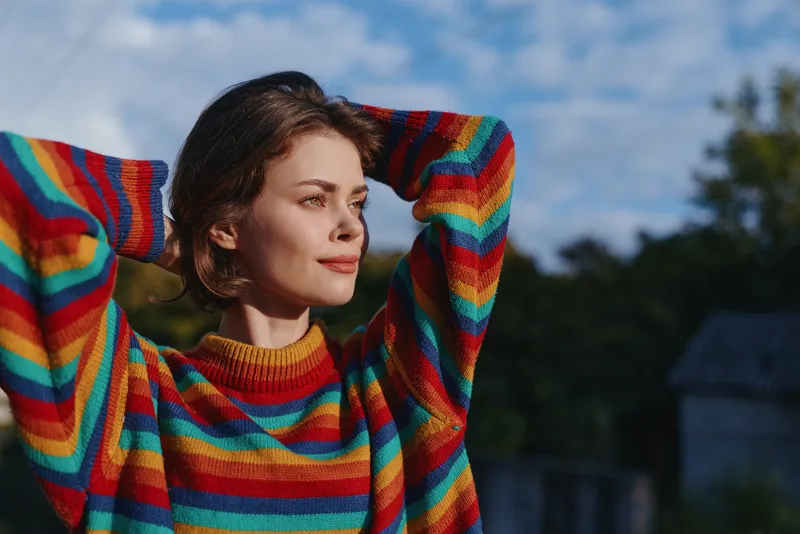 Woman, young woman, striped sweater, outdoors, smiling, casual outfit, hands behind head, looking away in a colorful knit on a sunny afternoon portrait for lifestyle, travel and cozy fashion.