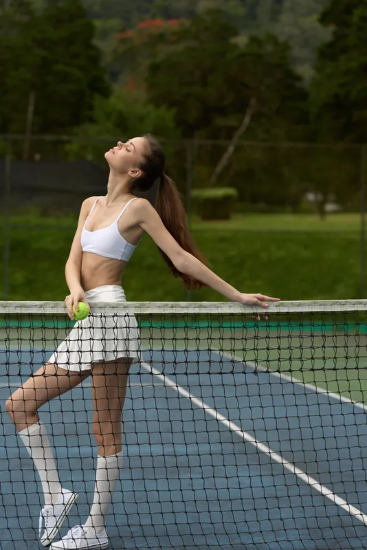 Young athletic model enjoying a moment of relaxation on a tennis court, wearing a white sports top and skirt, radiating confidence and joy amidst a lush green background