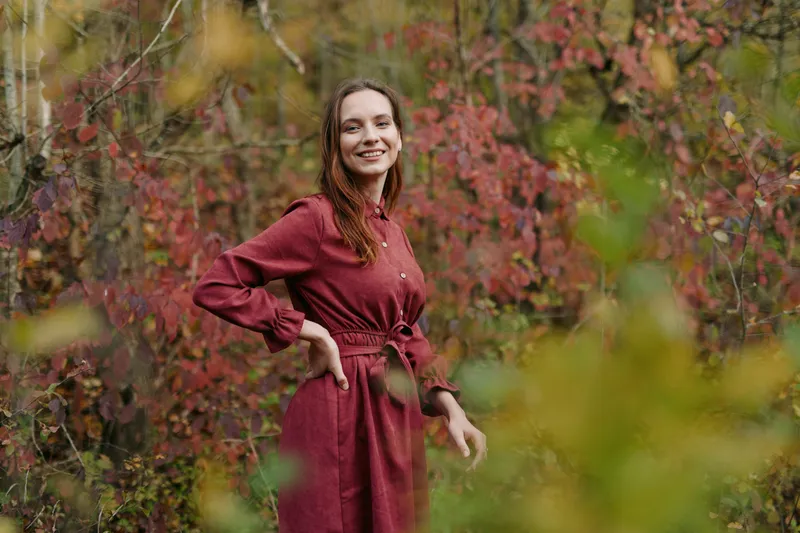 Young woman in a simple red dress smiles naturally among autumn foliage, her genuine expression and posture conveying authenticity and credibility in a candid outdoor portrait.