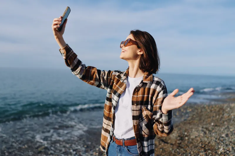 Woman taking a selfie on a sunny beach, wearing a plaid shirt and jeans, enjoying the sea breeze and ocean views with a raised arm.