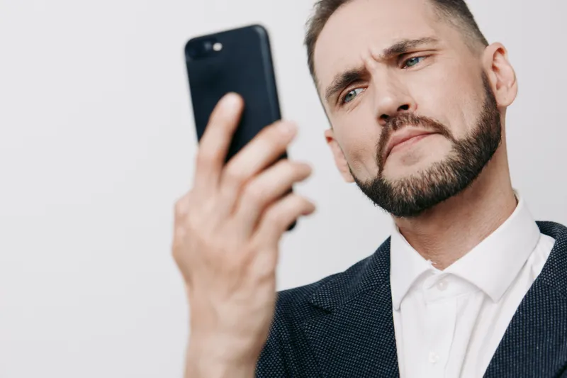 A professional man in a suit stands against a solid colored background, projecting a thoughtful emotion while checking his smartphone with calm confidence.