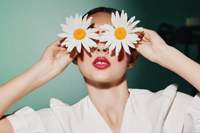 Young woman in white blouse holds two large white daisies over eyes, glossy pink lips and artful pose against muted teal background. Creative fashion and beauty concept with floral elements.