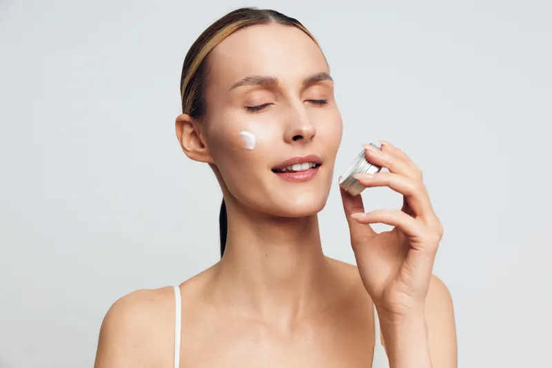 A young woman with clear skin applies face cream on her cheek with eyes closed in a bright studio. Concept for skincare, beauty routine, and self care.