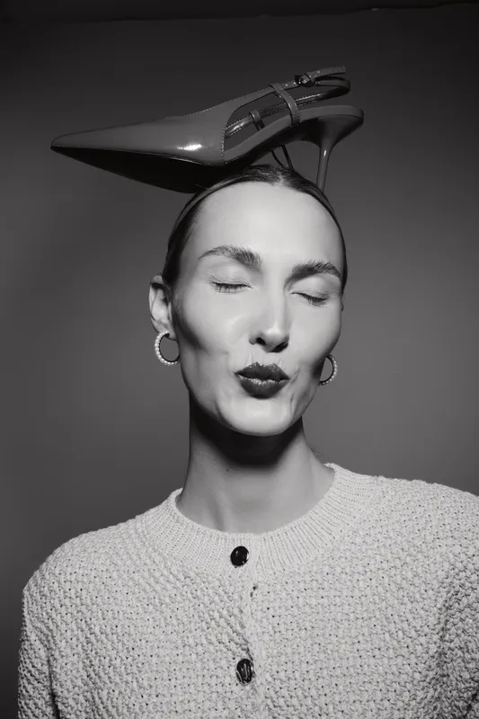 Elegant caucasian woman with light skin closes eyes and kisses with high heel shoe on head in black and white studio portrait.