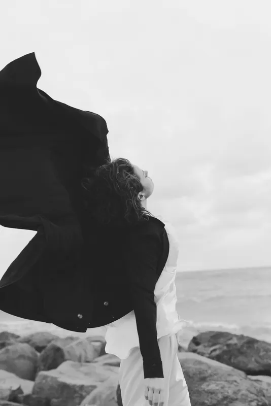 Embrace on a windy coast, a couple lifts a flowing cloak against the sea backdrop, black and white mood, expressive movement, intimate shoreline moment, fashion inspired