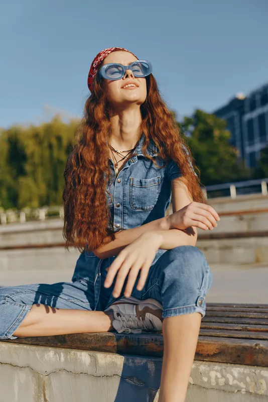 Lifestyle woman in modern boho-western denim street style sits outdoors on stairs wearing sunglasses and a headscarf, captured in warm film color tones.