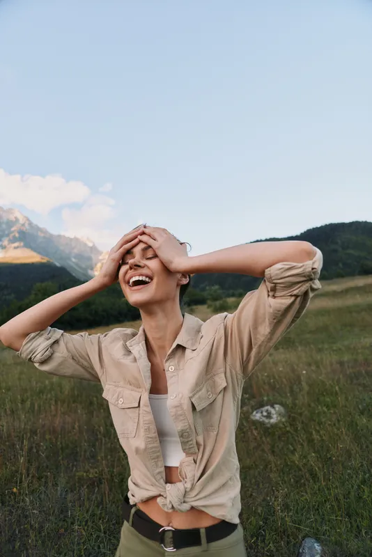 A young woman with brunette hair laughs in a sunny mountain meadow, wearing a beige tied shirt and white top, expressing carefree outdoor joy and freedom.