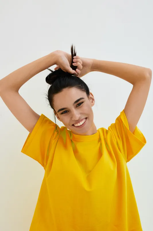 smiling woman in yellow t-shirt posing happily with arms raised against plain white background, expressing joy and casual style, natural light portrait