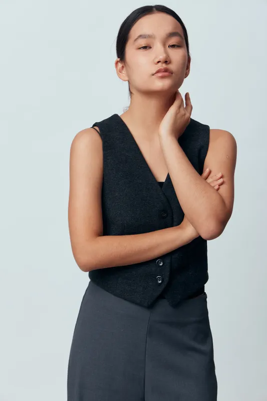 Young woman posing with thoughtful expression standing against light background wearing black sleeveless vest and dark skirt in studio. Confidence, fashion and portrait concept.