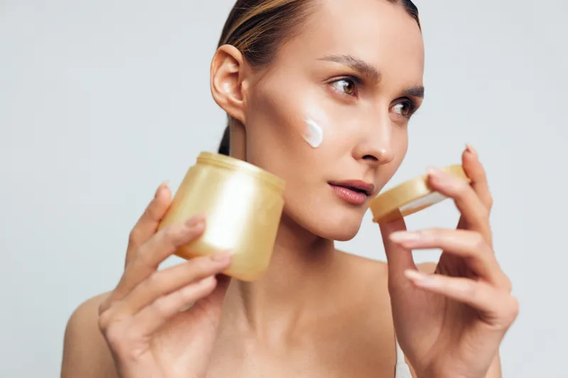 Woman applying white face cream, holding jar and small cup, close up on light gray studio lighting background. Skincare, wellness, self care, hydration, calm mood.