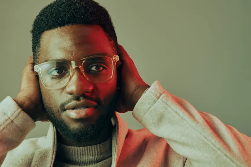 Young Black man with glasses showing apprehension, holding his head, in a softly lit modern indoor setting, expressing emotional depth and thoughtfulness