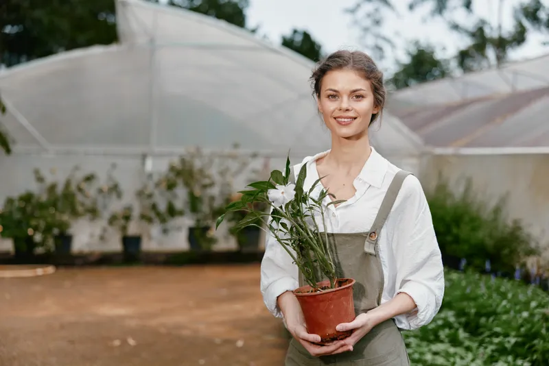 Young woman in overalls holding a potted plant in a greenhouse, smiling and surrounded by greenery, representing gardening and sustainable living