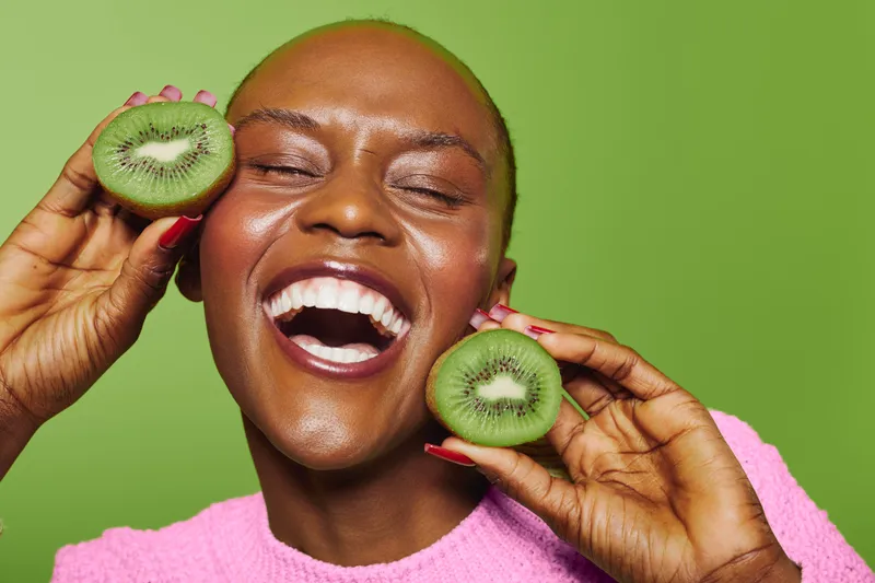 Happy dark skinned African woman with bald head smiling widely and holding two halves of kiwi fruit near face on green background. Concept of healthy lifestyle, fresh fruit, and joy.