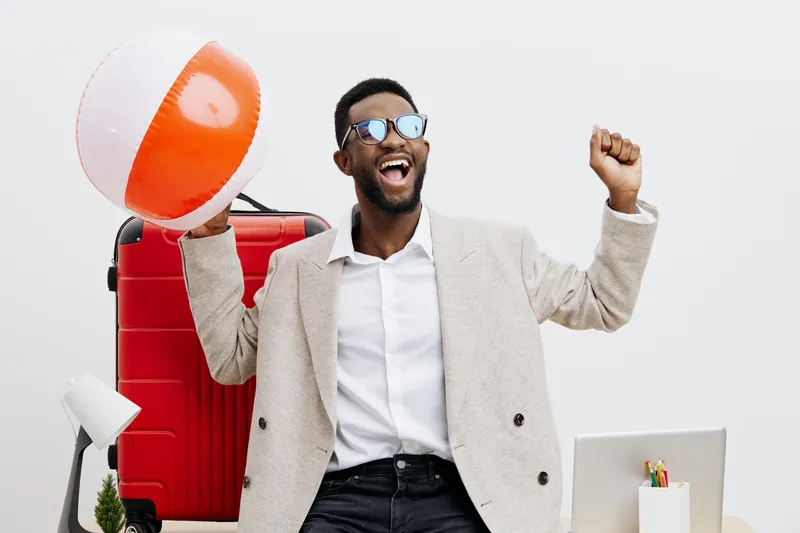 Smiling happy man with sunglasses holding beach ball in one hand and celebrating with fist pump in front of suitcase on plain white background. People lifestyle concept, vacation, holiday, summer fun.