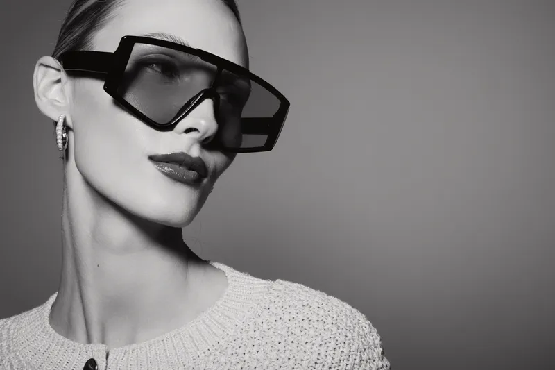 Stylish white woman with pale skin wearing oversized sunglasses and earrings poses in studio with textured sweater and confident expression.