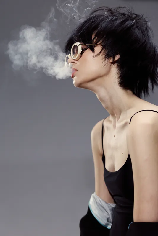 Stylish woman with short black hair wearing glasses blowing smoke against gray background.