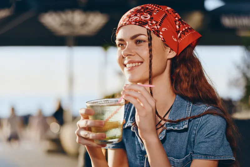 Smiling woman with red bandana enjoying cold drink outdoors in warm sunlight with blurred background, casual summer vibe and relaxed expression under clear sky.