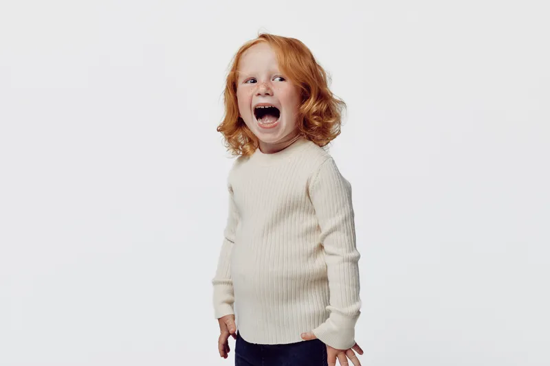 Любопытная рыжеволосая маленькая девочка стоит на белом фоне с открытым ртом