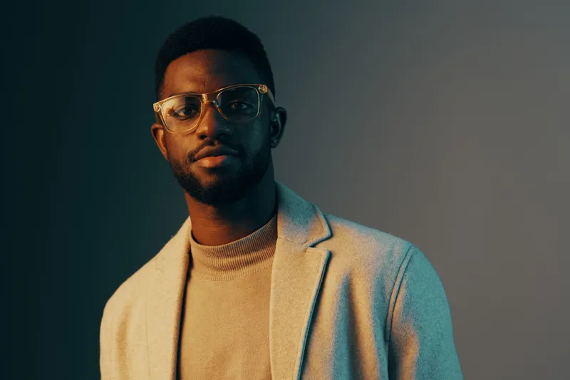 Stylish young Black man in glasses wearing a cozy sweater and light blazer, exuding confidence against a gradient background