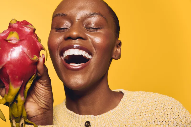 Happy African woman with dark skin laughs holding a dragon fruit near her face on yellow background. Concept of healthy lifestyle, exotic fruit, joy, and natural beauty.
