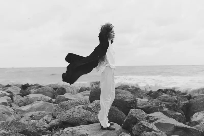 Woman stands on rocky shoreline in a flowing coat, windblown hair, gazing toward the sea. Monochrome scene captures motion, contrast, rugged coast, calm horizon, solitary presence, cinematic mood,
