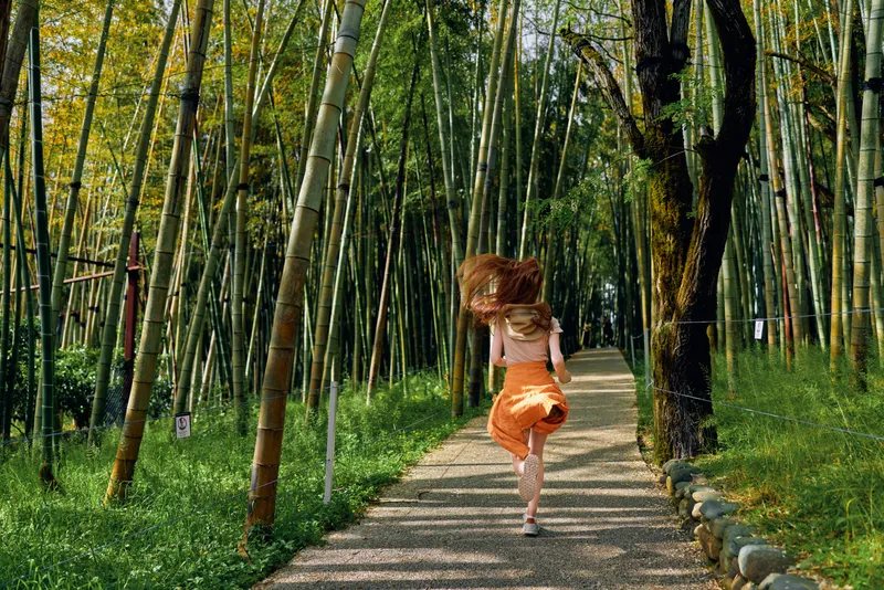 Girl running on a stone path through a dense bamboo forest wearing orange skirt, back view, motion and sunlight filtering through tall green stalks for outdoor nature walk.
