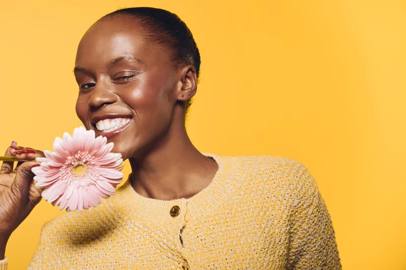 Smiling African American woman with dark skin holds pink flower near face on yellow background. Concept of happiness, beauty, natural skincare, and positive mood in studio lighting.