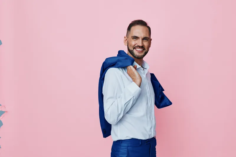 A confident businessman on a solid pink background, carrying a jacket over his shoulder, presenting a polished and professional attitude with calm confidence and friendly charm.