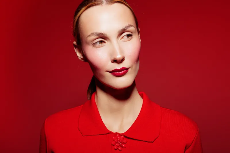 Portrait of a young woman in red sweater with red lipstick on red background. She looks to the side with a calm expression. Concept of fashion, beauty, and monochrome style.