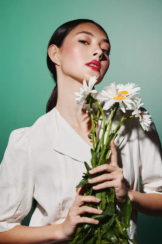 Elegant young woman with vivid red lipstick holding fresh daisies in hands, wearing soft white blouse against green background. Concept of beauty, fashion, and creative floral style.