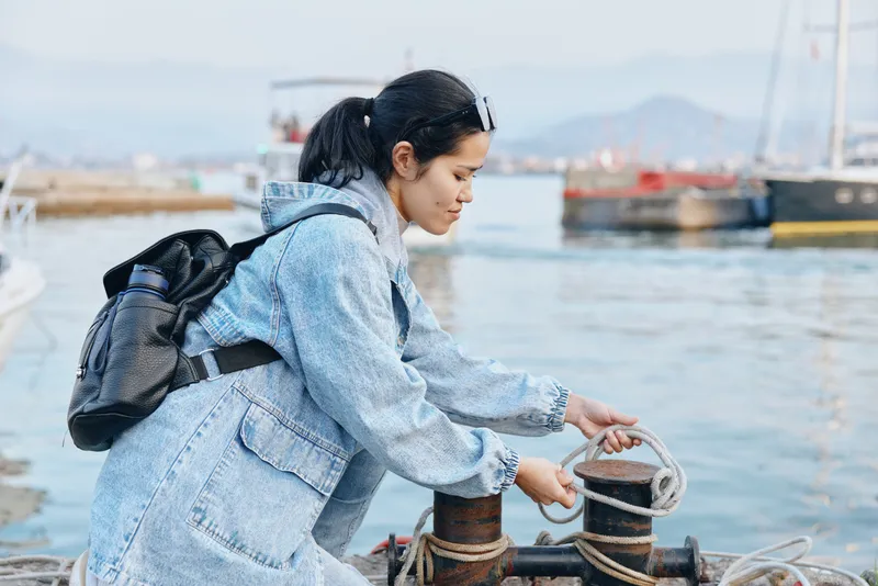 Harbor scene with a woman in a denim jacket tying rope to a bollard near moored boats, capturing a practical moment of outdoor maritime work and travel prep.