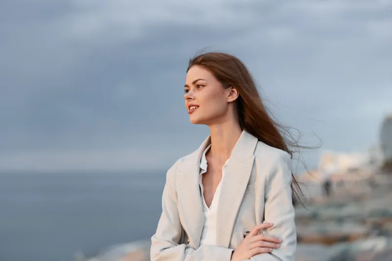 Young woman in light jacket looking thoughtfully over calm sea with cloudy sky, natural outdoor portrait conveying contemplation and peaceful mood