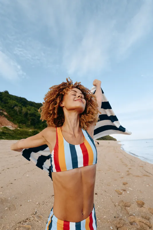 A young woman with curly hair in a striped swimsuit lifts her arms in a joyful, carefree pose on a sandy beach under a bright blue sky, with the ocean shimmering in the distance.