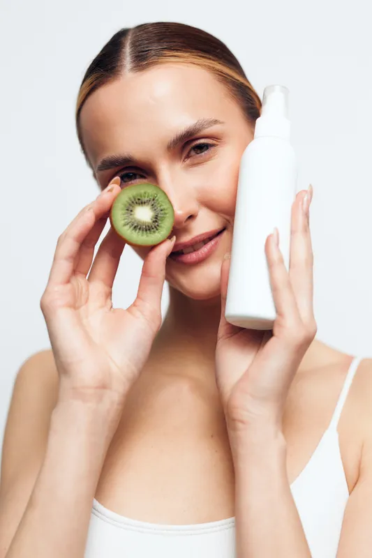 Woman skincare concept, holding a kiwi slice near her face and applying a clear serum bottle, smiling softly. Fresh natural beauty and health, modern self care.