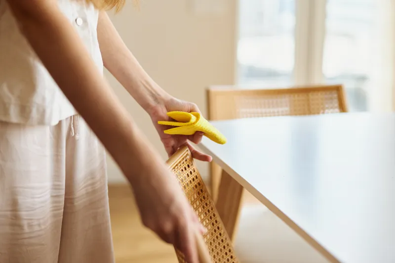 Cleaning woman wipes dining table and rattan chair with yellow sponge in a bright home, hands focused on surface care for household chores, tidy interior and daily maintenance.