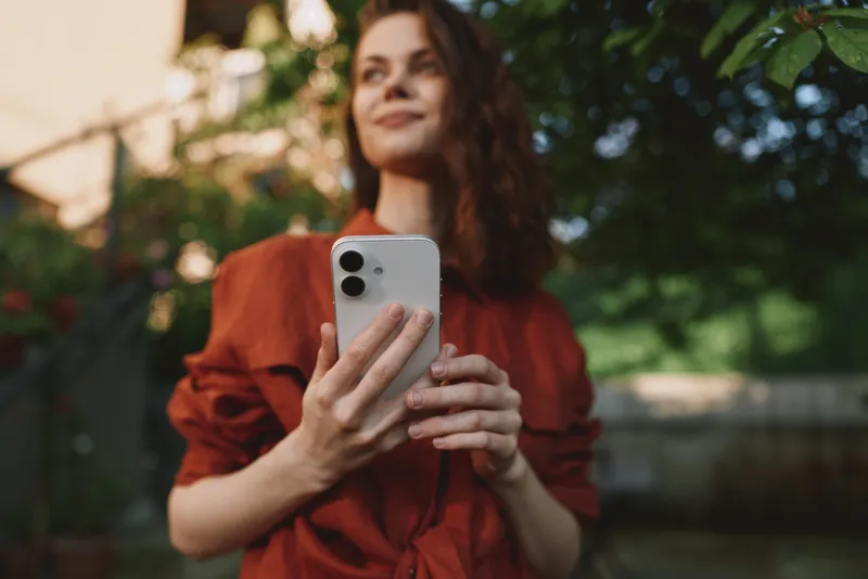 A young woman in a rust blouse holds a smartphone, capturing a candid moment outdoors with a relaxed, natural expression in a sunlit garden