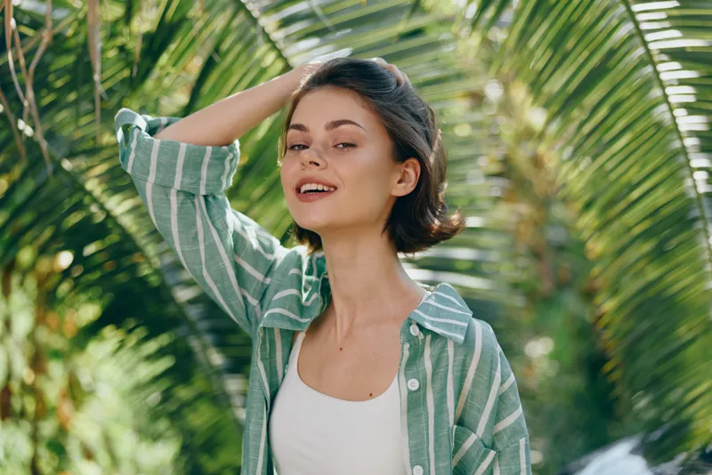 Bright portrait of a smiling woman outdoors among tropical palm leaves, wearing a green striped shirt and white top, bathed in warm sunlight.