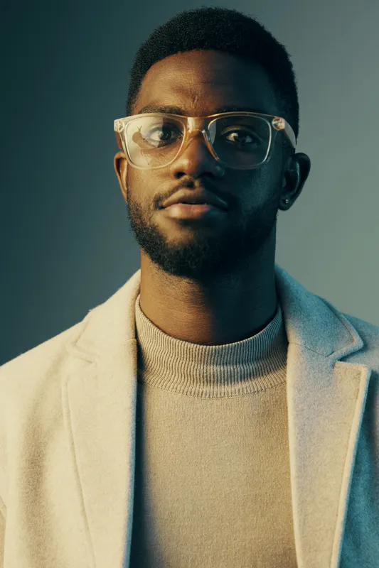 A confident young Black man wearing glasses and a stylish light colored coat poses against a gradient blue and green background, showcasing modern fashion and self assurance