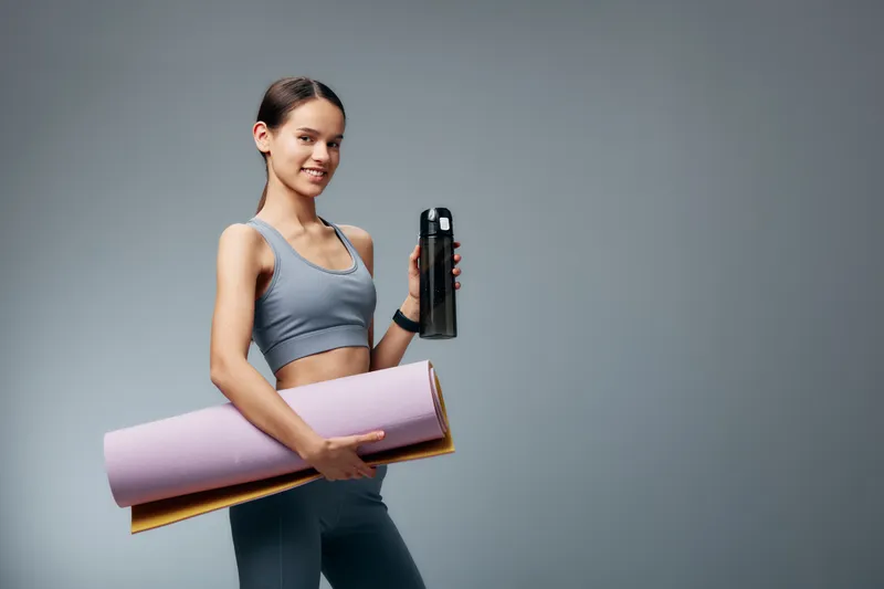 Fitness woman in athletic wear holds a water bottle and rolled yoga mat, ready for a workout. Studio backdrop, emphasizing healthy lifestyle, training routine and sport performance.