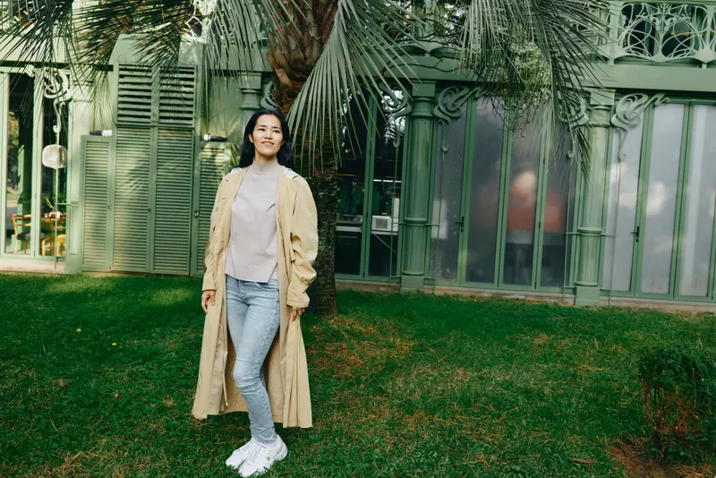 Woman wearing a beige trench coat, jeans and white sneakers stands on a green lawn beside a glass building, casual fashion and urban lifestyle captured in natural daylight.