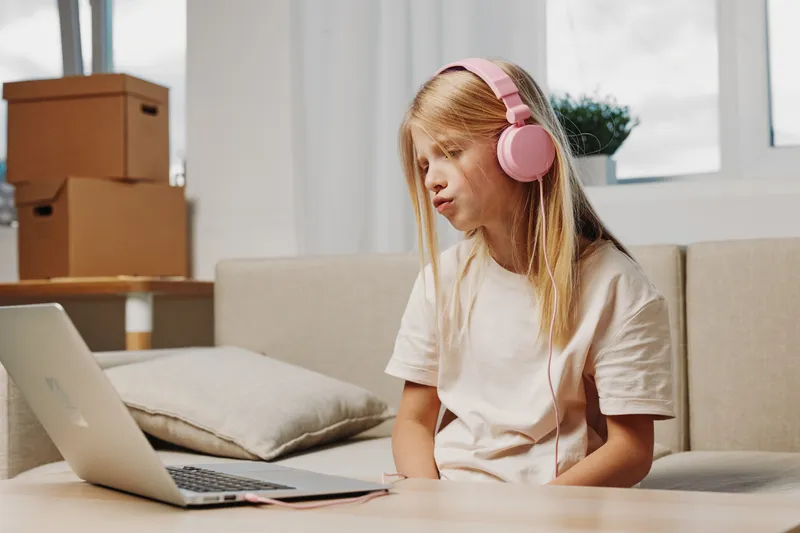 Young girl with long blonde hair wearing pink headphones watches laptop screen sitting on a beige sofa in cozy living room with natural light and cardboard boxes behind.