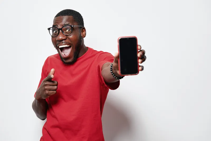 African American man with dark skin wearing glasses and red shirt showing smartphone with blank screen and pointing finger with excited expression on white background.