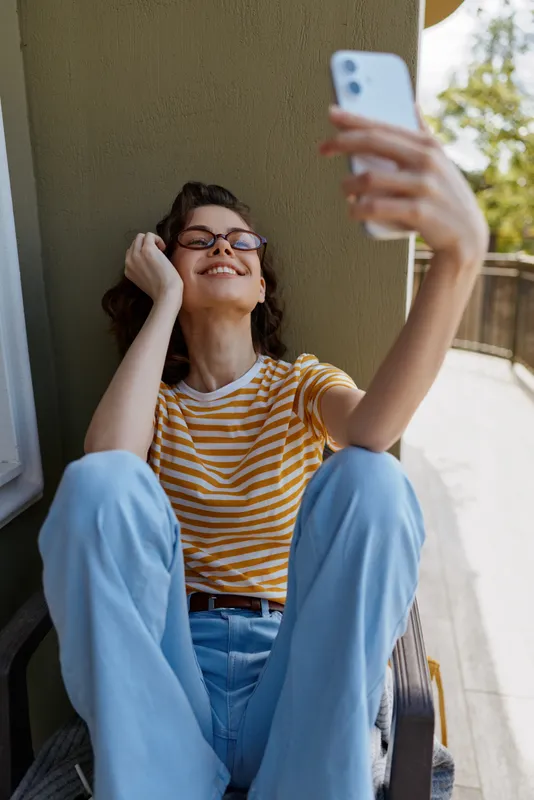 Smiling woman with glasses sits on a balcony step taking a selfie with a smartphone. Casual striped tee and jeans. Lifestyle moment highlighting confidence, self expression, and relaxed outdoor