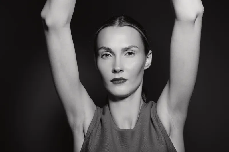 Portrait of a slim Caucasian woman with pale skin and dark hair, arms raised above head, wearing sleeveless top. Monochrome studio shot with dramatic light and shadow contrast.