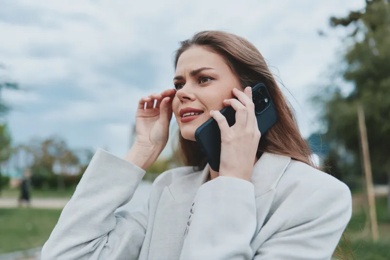 Professional businesswoman talking on the phone outdoors, expressing concern while wearing a light suit in a vibrant natural setting.