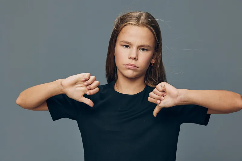Teenage girl with long hair in black t shirt showing dislike gesture with both thumbs down on grey background. Portrait of unhappy young female expressing disapproval.