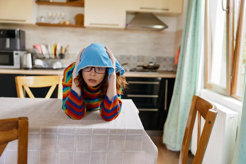 Young girl wearing a blue hat and colorful sweater rests her head on a kitchen table, a bright domestic scene that conveys cozy home life, playful mood, and casual everyday warmth.