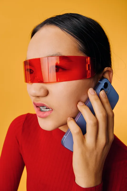 Woman with braces wearing red translucent visor talking on smartphone in studio closeup. Portrait of young female with serious expression in red outfit holding mobile device for communication.