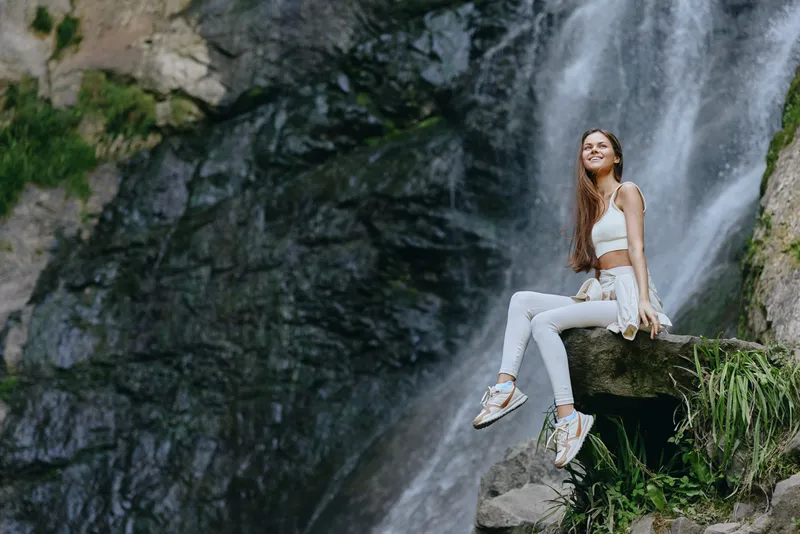 Young woman sitting on rock near waterfall, wearing white activewear and sneakers, with long hair and serene smile in lush outdoor nature setting.